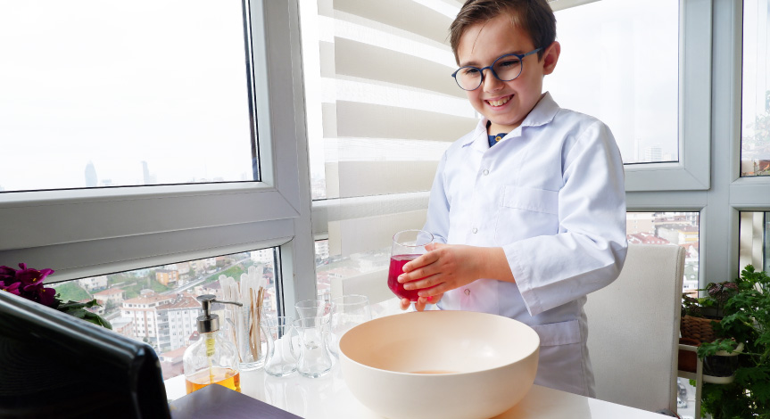 Boy in lab coat holding a glass with a red liquid standing in front of his computer performing a Mad Science virtual class.