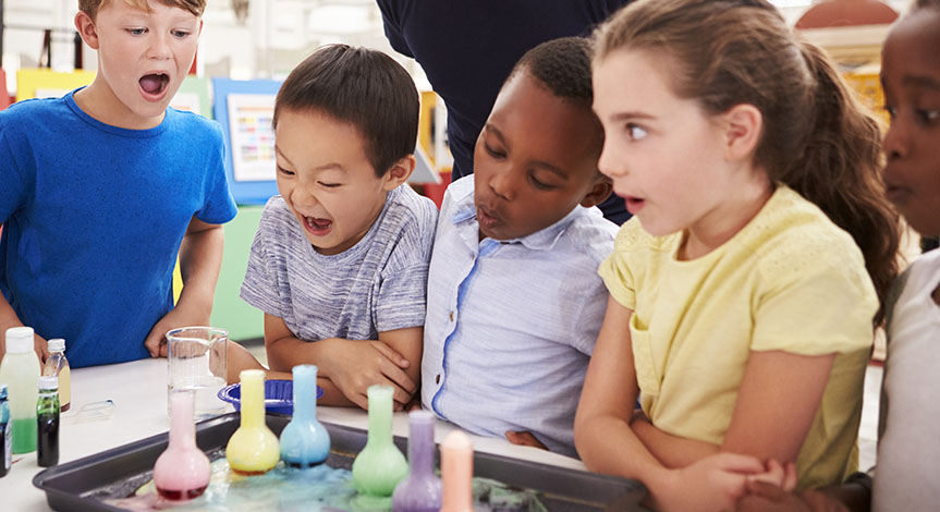 Teacher and kids watch experiment where they add colorful liquid into beakers