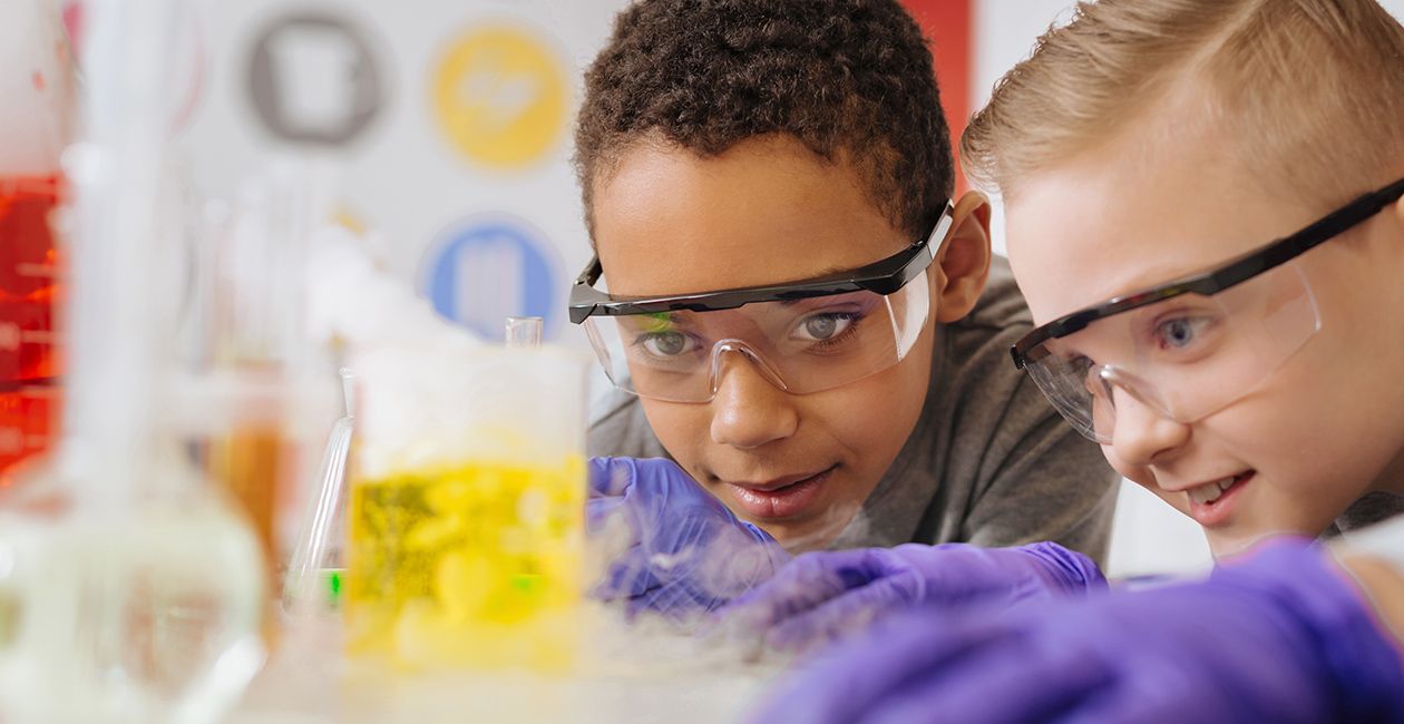 boys in safety goggles leaning closely to the table and watching chemical reaction going on in the beaker