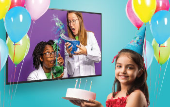 A girl holding a cake wearing a birthday hat watching a tv with a Mad Science instructors pouring a green liquid into a into