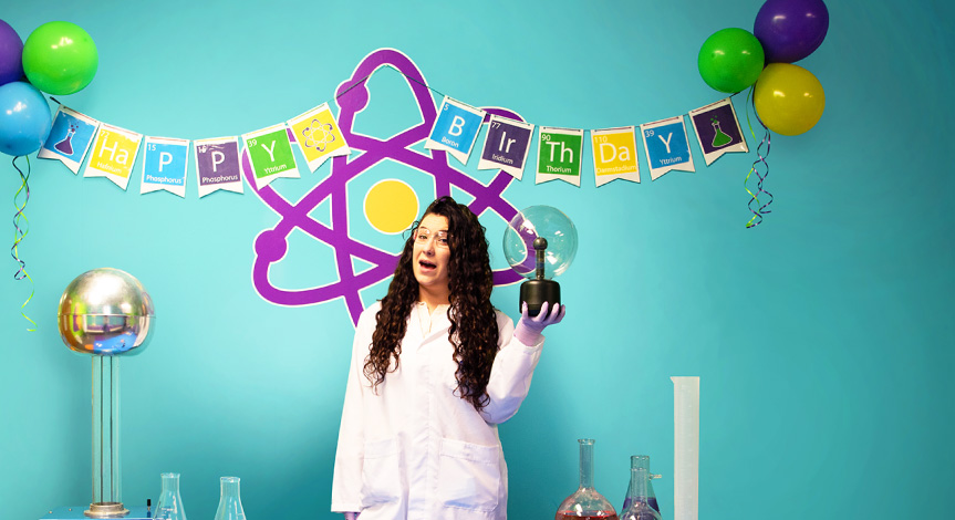 An instructor standing in front of an atom background with a plasma ball and beakers at a birthday party.