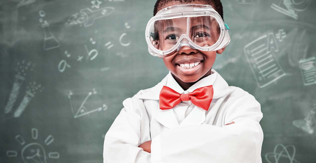 Boy holding a test tube filled with yellow liquid.  He is surrounded by other beakers and test tubes with multi colored liqui