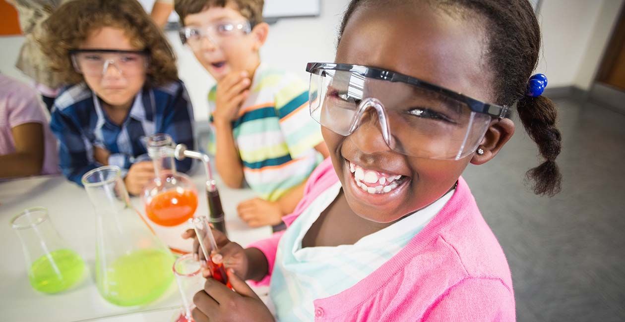 Girl smiling while holding a test tube with liquid inside. 2 other boys looking up at girl.  There are 2 beakers on the desk.